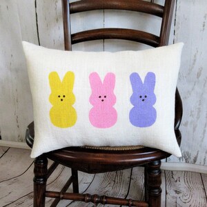 May include: A white cushion with three pastel-coloured bunny silhouettes. The bunnies are yellow, pink, and purple. The cushion is on a wooden chair.