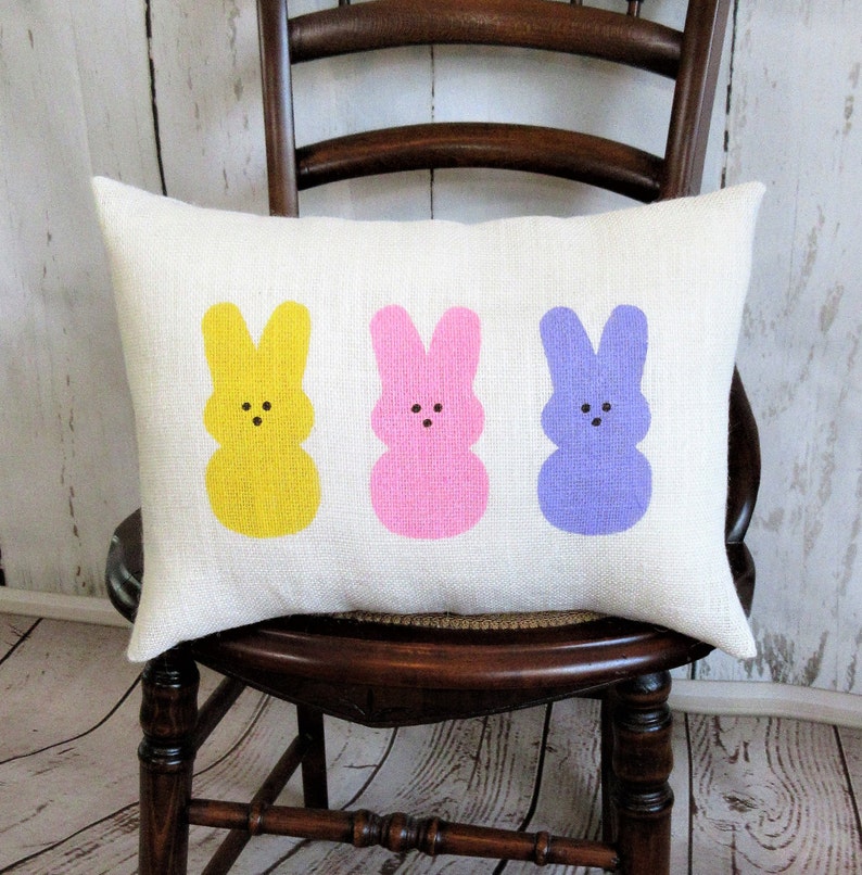 May include: A white cushion with three pastel-coloured bunny silhouettes. The bunnies are yellow, pink, and purple. The cushion is on a wooden chair.