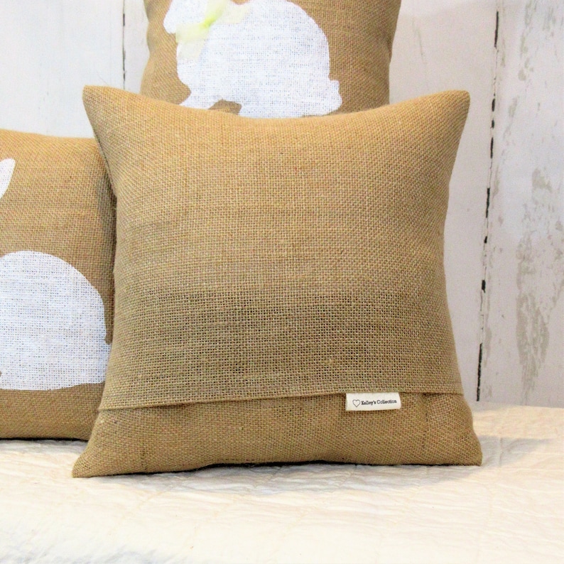 Puede incluir: Dos almohadas de tiro de arpillera con siluetas de conejos blancas. La almohada de arriba tiene una peque&ntilde;a etiqueta blanca con el texto "Bailey's Collection".