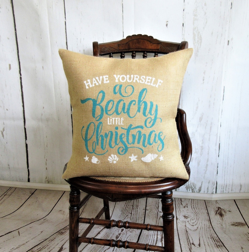 May include: A burlap pillow with the text "Have Yourself a Beachy Little Christmas" in teal blue lettering with white accents. The pillow is sitting on a wooden chair.