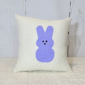 May include: A white burlap pillow with a purple bunny rabbit design. The bunny has black eyes and a simple, smiling face.