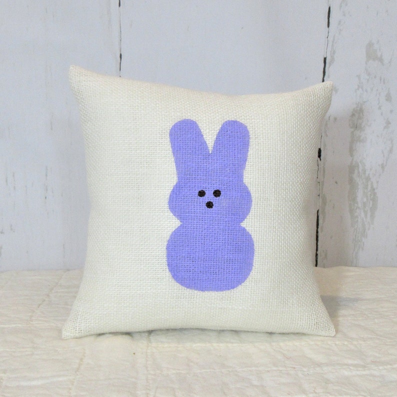 May include: A white burlap pillow with a purple bunny rabbit design. The bunny has black eyes and a simple, smiling face.