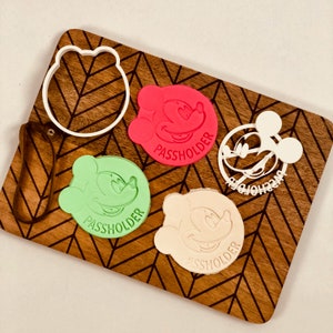 May include: Four cookie cutters featuring Mickey Mouse's head with the text "Passholder" on each. The cutters are in red, green, white, and a light brown color. A white cookie cutter with Mickey Mouse's ears is also included.
