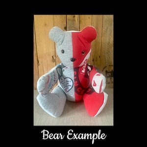 May include: A handmade teddy bear with a split design. One side is gray, the other is red with white text. The bear has a heart-shaped patch with text. The bear is sitting on a light-colored surface.