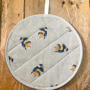 May include: Circular, quilted pot holder featuring a light grey fabric with a pattern of yellow, black, and orange watercolour bees. It has a white fabric loop for hanging. The diameter is approximately 20 cm.