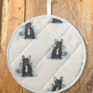 May include: Circular, quilted pot stand with a repeating pattern of black and white French Bulldogs. The pot stand has a light grey background with a stitched diamond pattern and a light grey trim. A white fabric loop is attached for hanging.