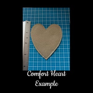 May include: A tan heart-shaped felt patch with a silver border, approximately 3 inches in height, is displayed on a blue grid surface. The text "Comfort Heart Example" is written in cursive below the heart.