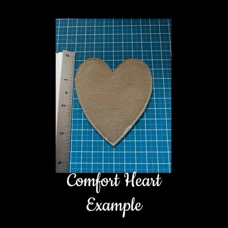 May include: A tan heart-shaped felt patch with a silver border, approximately 3 inches in height, is displayed on a blue grid surface. The text "Comfort Heart Example" is written in cursive below the heart.