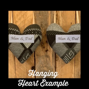 May include: Two heart-shaped fabric ornaments with a gray and green argyle pattern. Each heart has a white label with the embroidered text "Mum & Dad". The hearts hang from a cream-colored ribbon against a wooden background.