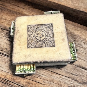 May include: A ceramic matchbox with a sun and moon design on the front. The box has four sides, each with a drawer that holds matches. The matches are green and white.