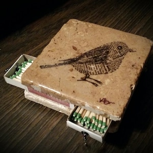 May include: A small, square, beige tile box with a drawer that opens to reveal a row of matches. The tile is decorated with a black and white image of a bird with a long tail.