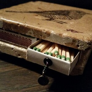 May include: A small, rectangular matchbox made of stone with a bird design on the top. The matchbox has a drawer that pulls out to reveal a full box of matches.