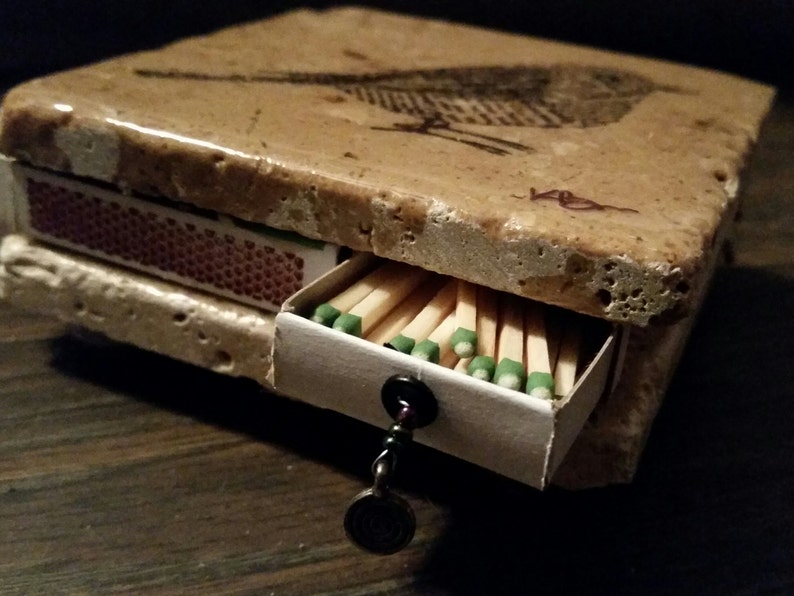 May include: A small, rectangular matchbox made of stone with a bird design on the top. The matchbox has a drawer that pulls out to reveal a full box of matches.