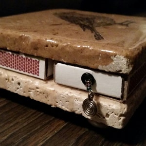 May include: A small, decorative box made of stone with a bird design on the top. The box has a drawer and a small compartment on the side. The box is decorated with a silver chain and a spiral charm.