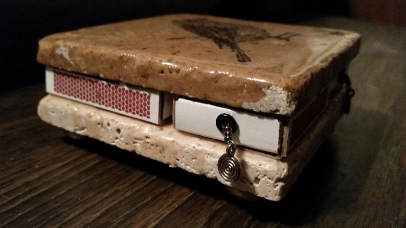 May include: A small, decorative box made of stone with a bird design on the top. The box has a drawer and a small compartment on the side. The box is decorated with a silver chain and a spiral charm.
