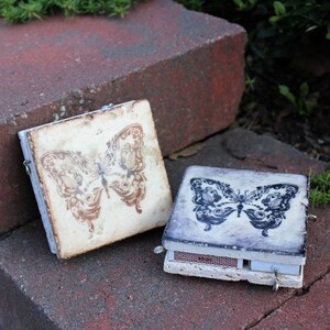May include: Two small, square boxes with butterfly designs. The box on the left has a brown butterfly on a white background. The box on the right has a black butterfly on a gray background. Both boxes have a small metal ring attached.