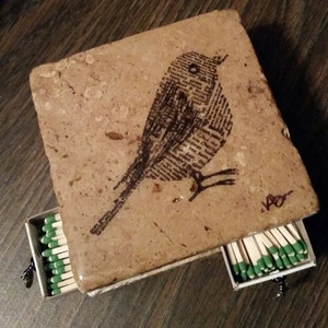 May include: A small, rectangular, brown stone box with a drawer that slides out. The box has a black and white image of a bird printed on the top. The drawer is open and contains a row of matches with green tips.