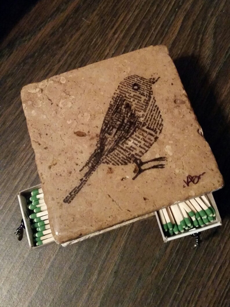 May include: A small, rectangular, brown stone box with a drawer that slides out. The box has a black and white image of a bird printed on the top. The drawer is open and contains a row of matches with green tips.