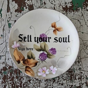 Op de afbeelding: Een wit keramiek bord met een handgeschilderd ontwerp van een wijnstok met paarse bessen en roze bloemen. Het bord heeft de tekst "Sell your soul" en "Sign here:" in zwart.