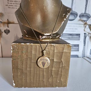 May include: A vintage gold-toned locket necklace displayed on a gold-painted bust. The round locket features an ornate design with a central, stylized shape. The necklace chain is delicate and complements the antique aesthetic.