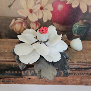 Puede incluir: Una escultura de flor de jade blanco con cuentas rojas en el centro, descansando sobre una caja de madera con el texto "AMERICAN CIGARS" impreso en el lateral.