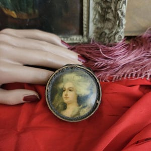May include: A small, round, wooden box with a painted portrait of a woman wearing a white wig and a blue dress. The box is on a red fabric surface.