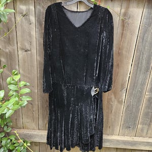 May include: A black velvet dress featuring a V-neckline, long sleeves, and a subtle tonal wood grain pattern. The garment includes a drop-waist silhouette with a side-gathered skirt detail accented by a metallic buckle on the right hip.