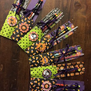 May include: A collection of seven Halloween-themed gift sets, each featuring decorative paper tags with patterns of pumpkins, candy corn, and cats. Each set includes pencils tied with purple ribbon and adorned with layered paper flower embellishments.