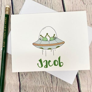 May include: A white card with a green alien spaceship and the name Jacob written in green letters.