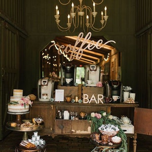 May include: A rustic coffee bar setup with a wooden sign that says "coffee" and a sign that says "bar". There are two coffee makers, a cake, and various coffee supplies on a wooden table. The coffee bar is decorated with greenery and flowers.