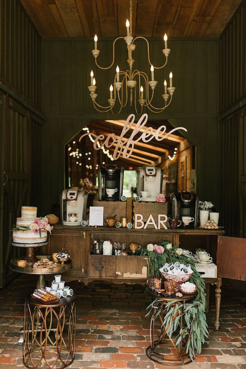 May include: A rustic coffee bar setup with a wooden sign that says "coffee" and a sign that says "bar". There are two coffee makers, a cake, and various coffee supplies on a wooden table. The coffee bar is decorated with greenery and flowers.