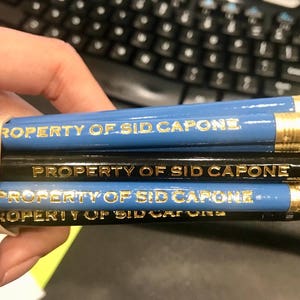 May include: A close-up of a stack of blue and black pencils with gold lettering that reads "PROPERTY OF SID CAPONE".