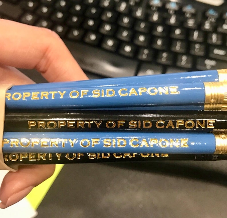 May include: A close-up of a stack of blue and black pencils with gold lettering that reads "PROPERTY OF SID CAPONE".