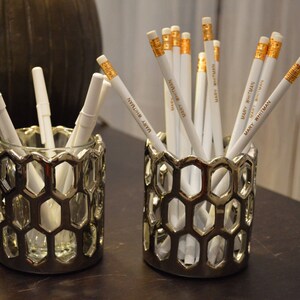 May include: Two silver hexagonal pencil holders with white pencils inside. The pencils have gold lettering that says "Mary Whitman" and "Stephanie Ashton".