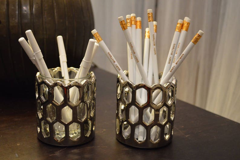 May include: Two silver hexagonal pencil holders with white pencils inside. The pencils have gold lettering that says "Mary Whitman" and "Stephanie Ashton".