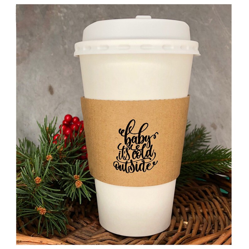 May include: White takeaway coffee cup with a brown paper sleeve. The sleeve has black text that reads "Baby, it's cold outside". The cup is sitting on a woven wicker surface with a green pine branch and red berries.