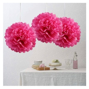 May include: Three large pink tissue paper pom-poms hanging from the ceiling above a table set for a party.