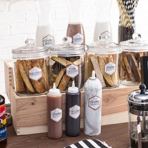 May include: A coffee bar set up with a variety of syrups, toppings, and biscotti. The syrups are in clear glass bottles with gold pumps and labels that say "Torani". The toppings are in clear glass jars with labels that say "Biscotti", "Dark Chocolate", "Lemon", and "Whipped Cream". The biscotti are in clear glass jars with labels that say "Biscotti", "Dark Chocolate", "Lemon", and "Whipped Cream". The sign says "Everything I Brew, I Brew For You".