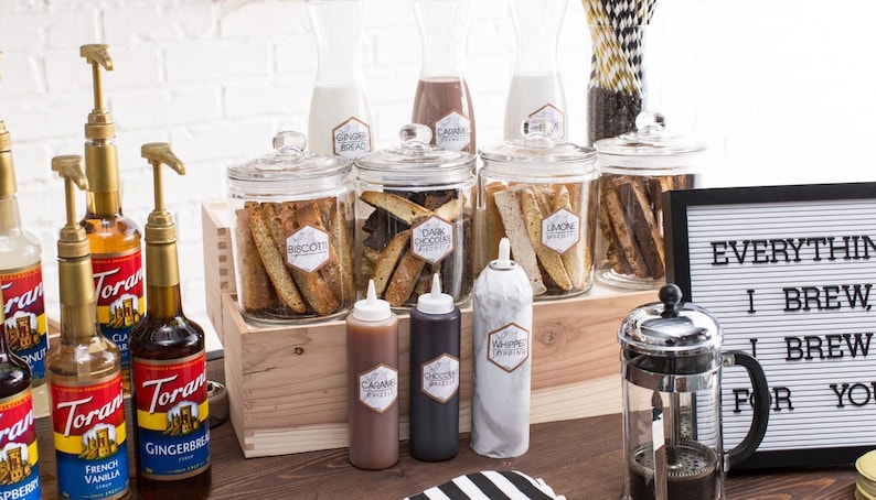 May include: A coffee bar set up with a variety of syrups, toppings, and biscotti. The syrups are in clear glass bottles with gold pumps and labels that say "Torani". The toppings are in clear glass jars with labels that say "Biscotti", "Dark Chocolate", "Lemon", and "Whipped Cream". The biscotti are in clear glass jars with labels that say "Biscotti", "Dark Chocolate", "Lemon", and "Whipped Cream". The sign says "Everything I Brew, I Brew For You".