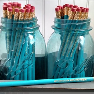 May include: Two blue glass jars filled with teal pencils. The pencils are labeled "Studio on Seventeen".