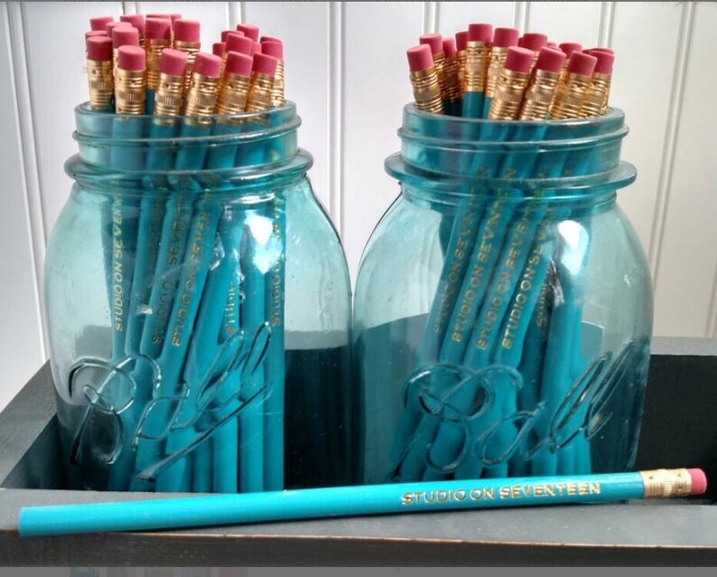 May include: Two blue glass jars filled with teal pencils. The pencils are labeled "Studio on Seventeen".