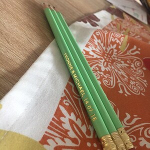 May include: Two green pencils with gold lettering that reads "Yvonne & Michael 14.07.18" on a patterned fabric.