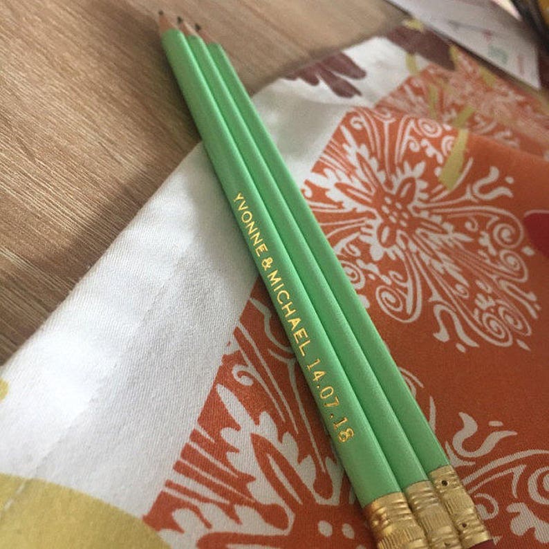 May include: Two green pencils with gold lettering that reads "Yvonne & Michael 14.07.18" on a patterned fabric.