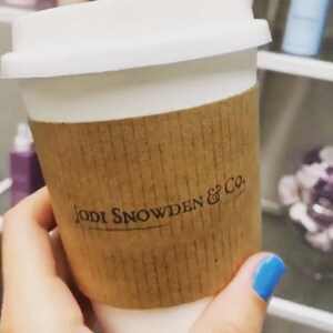 May include: A white takeaway coffee cup with a brown paper sleeve. The sleeve has the text "JODI SNOWDEN & CO." printed on it.