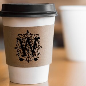 May include: A white takeaway coffee cup with a black lid and a brown paper sleeve. The sleeve features a black and white logo with a stylised letter "W" surrounded by decorative flourishes.