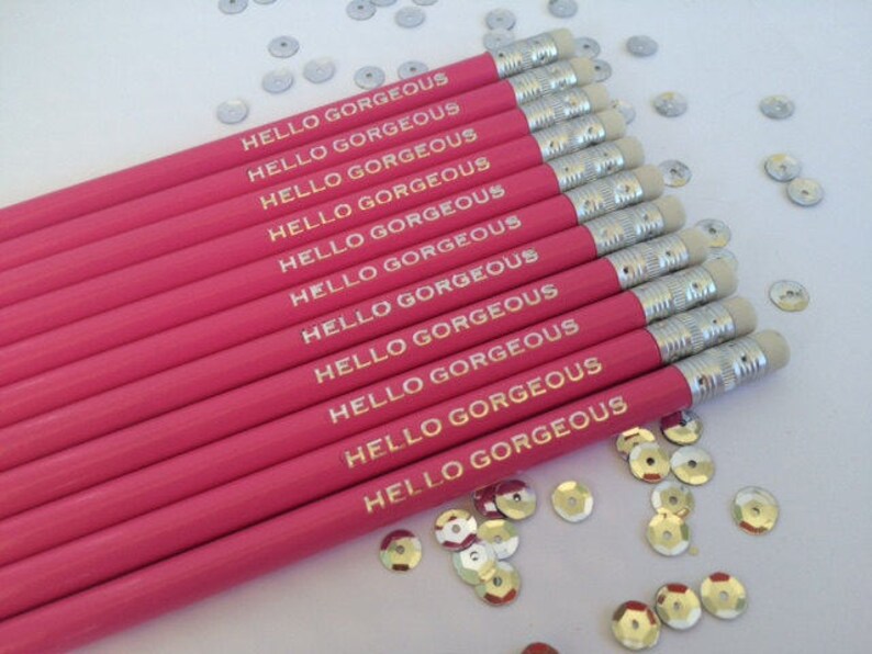 May include: A set of pink pencils with gold lettering that reads "Hello Gorgeous" on each pencil. The pencils are arranged in a stack with the erasers facing up.