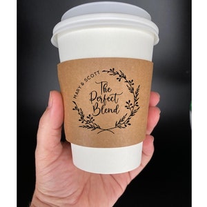 May include: A white coffee cup with a brown paper sleeve. The sleeve has a black wreath with the text "The Perfect Blend" and "Mary & Scott" on it.