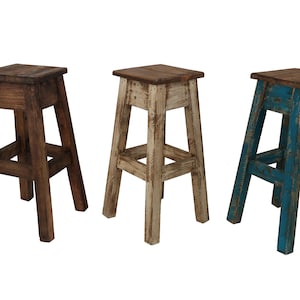 May include: Three rustic wooden stools. One is dark brown, one is off-white, and the third is blue. Each stool has a square seat and angled legs, showcasing a distressed finish. The stools are suitable for a kitchen or bar.