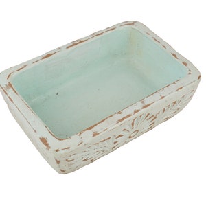 Pu&ograve; includere: Un vaso rettangolare in ceramica con una finitura bianca usurata e un interno blu chiaro. Il vaso ha un motivo floreale attorno al bordo.