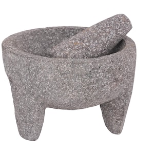 May include: A gray stone mortar and pestle set. The mortar has a rounded bowl shape with three sturdy legs. The pestle rests inside the bowl. The set is made of a speckled, textured stone.